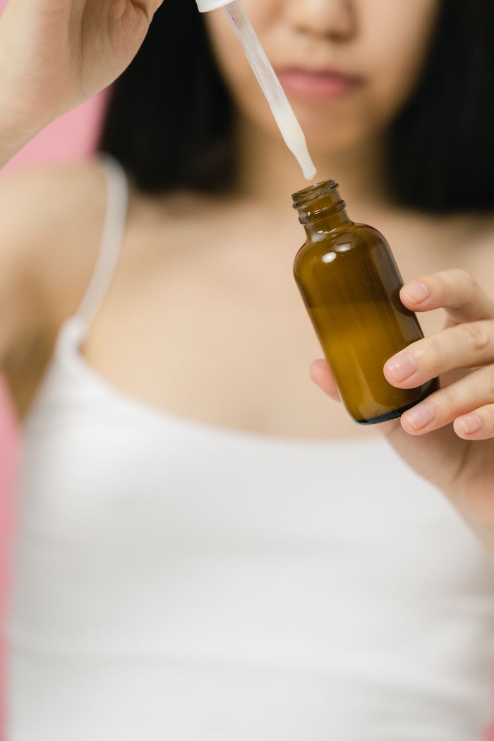 A woman in a white tank top applies serum from a dropper to a glass bottle, highlighting skincare.